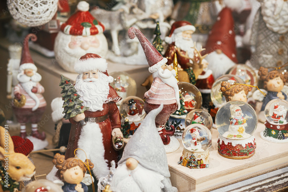 Stylish Christmas souvenirs, santa clauses, snow globes, snowman toys in showcase of festive store. Modern christmas decor in city street. Winter holidays in Europe. Merry Christmas