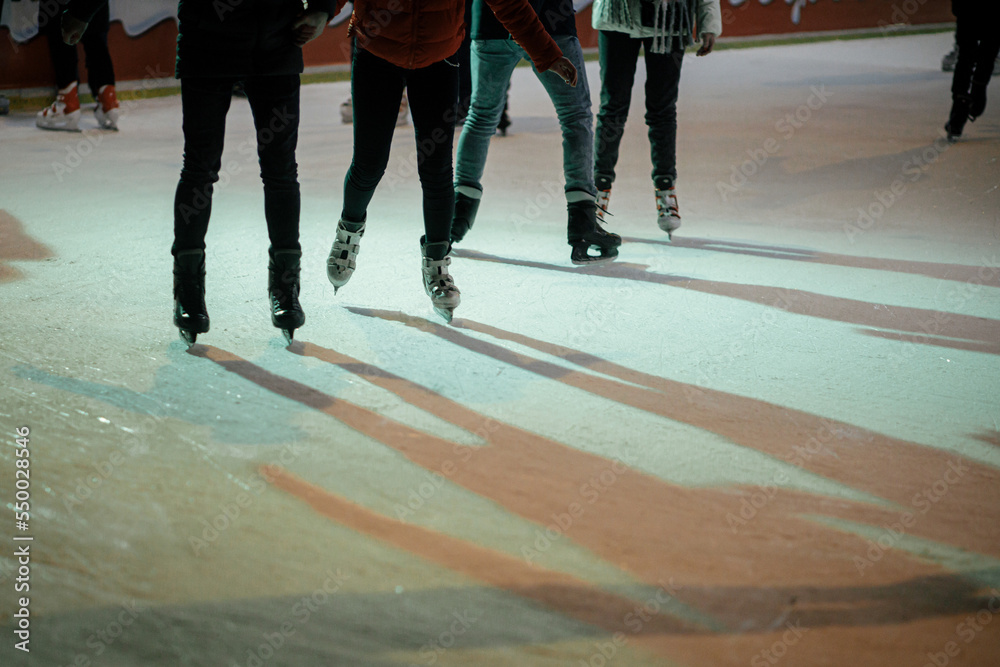 Obraz premium Ice skaters on ice rink, skates close up. People having fun and skating in town square in evening.Legs with ice skaters cropped view. Atmospheric Winter holidays in Europe. Happy Holidays!