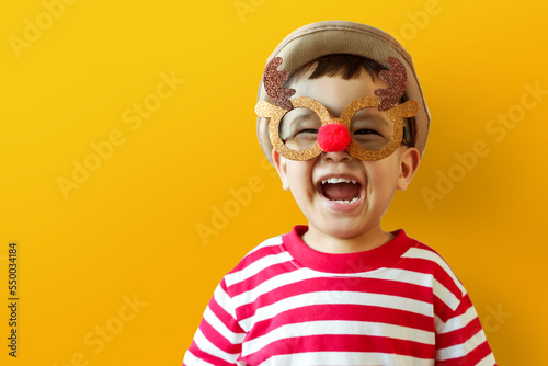 Happy funny Little Asian child boy in Christmas reindeer glasses and hat laughing on yellow isolated background.