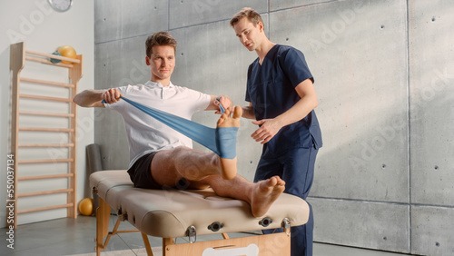 Young Male Athlete Undergoing Physiotherapy, Professional Sport Masseur Helping with Foot Exercise with a Rubber Band. Musculoskeletal Pain Therapy and Rehabilitation Concept.