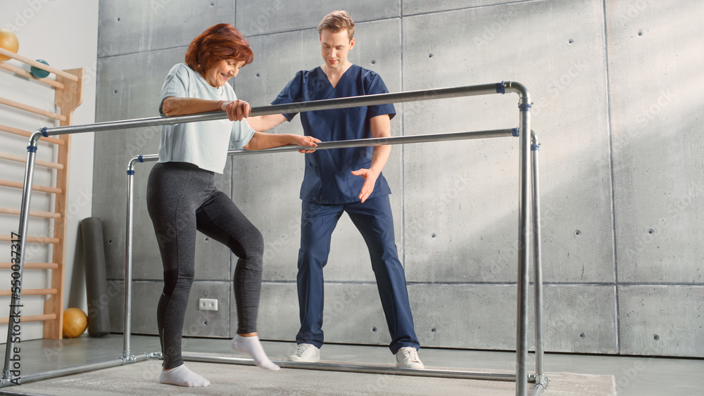 Fototapeta premium Physical Therapy Center: Portrait of Strong Senior Woman with Leg Injury Successfully Walks Holding Parallel Bars. Physiotherapist, Rehabilitation Doctor, Trauma Prevention Therapy Hospital.
