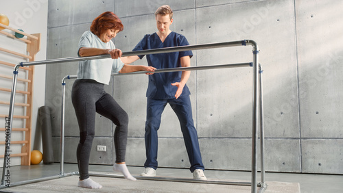 Physical Therapy Center: Portrait of Strong Senior Woman with Leg Injury Successfully Walks Holding Parallel Bars. Physiotherapist, Rehabilitation Doctor, Trauma Prevention Therapy Hospital.