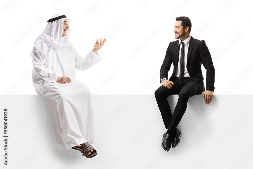 Arab man in ethnic clothes talking to a businessman seated on a blank white panel Arab man in ethnic clothes talking to a businessman seated on a blank white panel