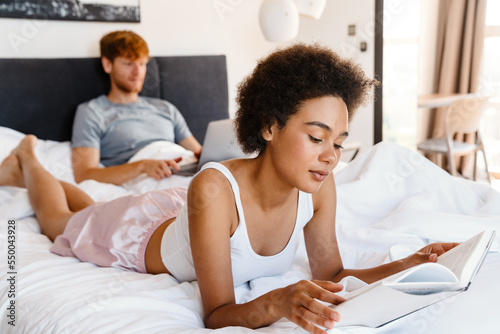 Young beautiful african woman reading lying with her working boyfriend photo