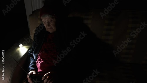 Ukraine. Grandmother holds radio in hands sitting in darkness blackout during russian terroristic attacks. Senior woman in winter clothes in darkness on sofa with hand radio receiver listening news. photo