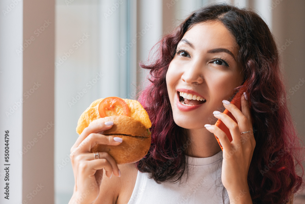 A woman eats a hamburger in a fast food cafe and talks to a friend on the phone A woman eats a hamburger in a fast food cafe and talks to a friend on the phone