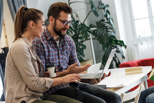 Two casually dressed young businesspeople at the office. They're sitting on a desk and discussing new ideas while drinks coffee.	