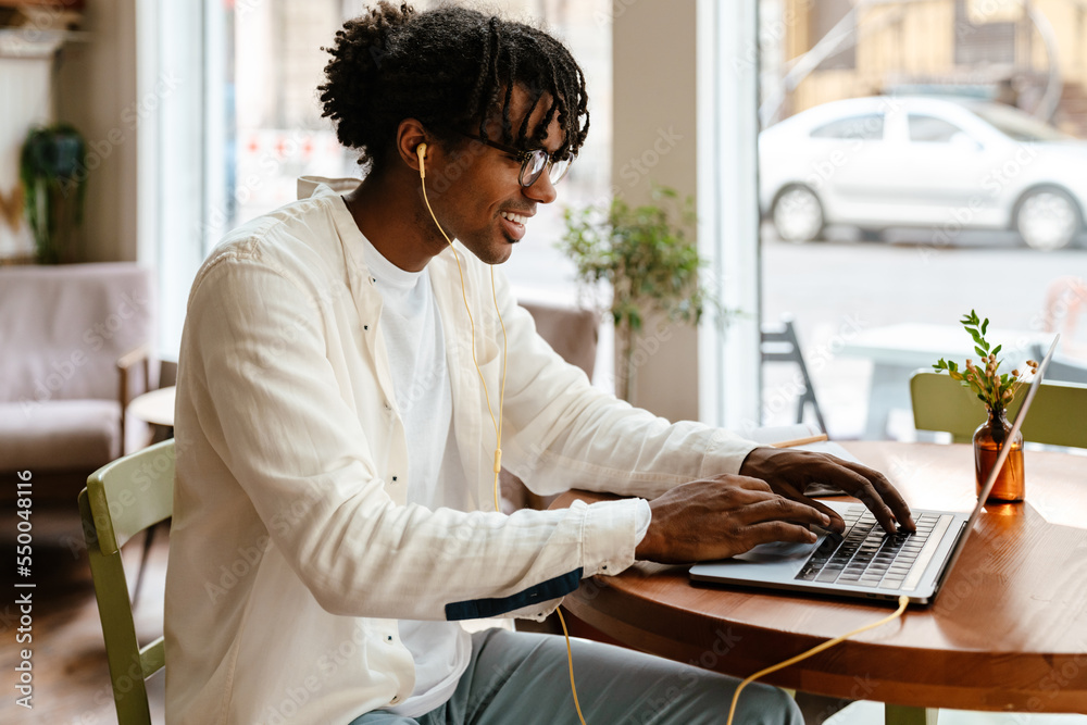 Young african american man using laptop and earphones in cafe indoors Young african american man using laptop and earphones in cafe indoors