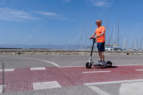 Uomo anziano con una maglia arancione corre sul suo monopattino elettrico  in una pista ciclabile  