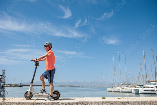 Uomo anziano con una maglia arancione e caschetto protettivo , corre sul suo monopattino elettrico di fronte al porto turistico ., isolato su sfondo cielo . 
