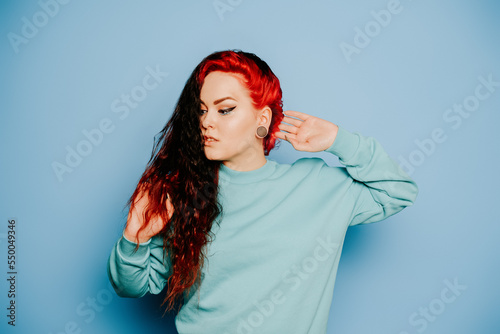 Woman in a blue sweater on a blue background posing. A beautiful brunette with long hair and and tunnels in her ears. Eyeliner and trendy makeup. © MoreThanProd