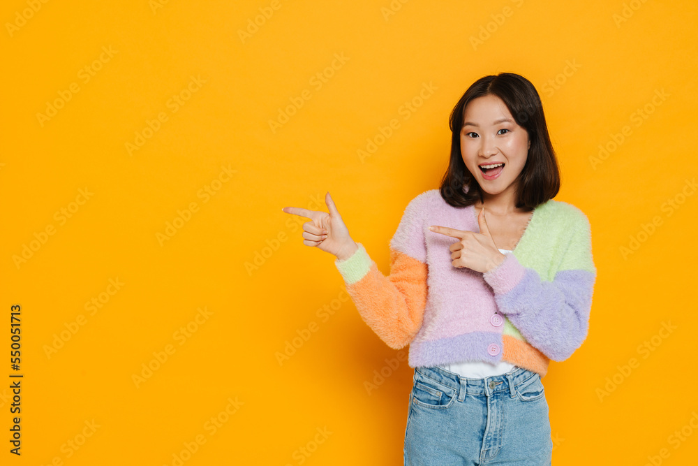 Young asian girl smiling and pointing fingers at copy space isolated over yellow background Young asian girl smiling and pointing fingers at copy space isolated over yellow background