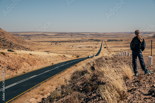 Ein Mädchen schaut am Straßenrand entlang einer Straße auf zwei entgegenkommende Autos - die Straße verliert sich in der endlosen Weite der Wüste (Aus, Namibia) photo