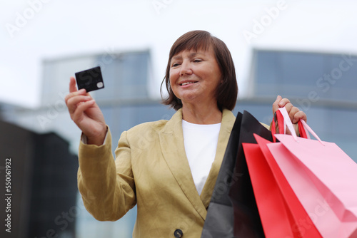 Smiling stylish middle aged woman with credit, debit card and shopping bags standing on shopping mall background. Autumn holidays, discounts, black Friday. concept of consumerism, sale, rich life.