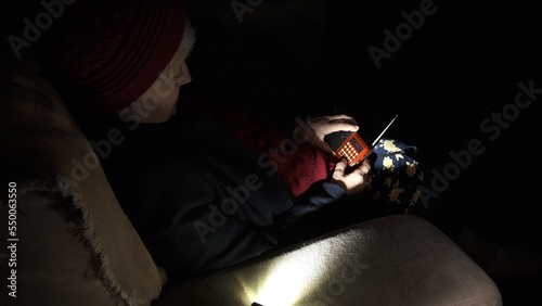 Ukraine: Grandmother holds radio in hands sitting in darkness blackout during russian terroristic attacks. Senior woman in winter clothes in darkness on sofa with hand radio receiver listening news