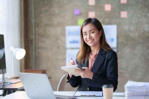 Beautiful Asian businesswoman working on paperwork at her desk in the office review work from different department, take minute of meetings report other document.