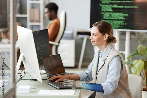 Smiling woman with bionic arm coding on computer when working on big project at research center photo