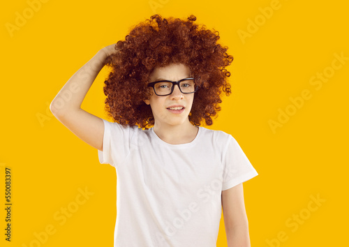 Portrait of funny puzzled child. School boy in curly brown wig and eyeglasses standing isolated on yellow background, thinking, scratching head and looking at camera with confused, unsure expression