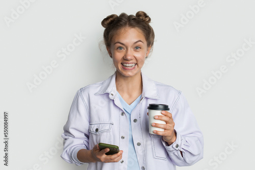 Young smiling happy student holding paper cup of coffee using mobile cell phone surfing internet, messaging friends online isolated on white background studio