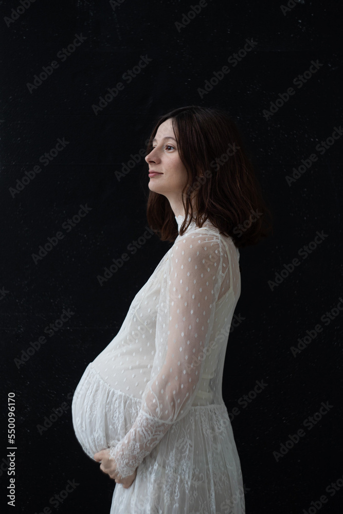 Fototapeta premium a pregnant brunette stands in profile on a black background in a white lace wedding dress hugs her big belly and looks away