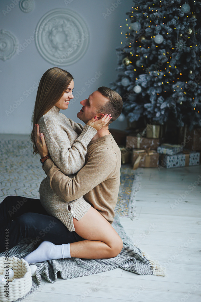 A couple in love on New Year's Eve in a cozy home environment near the Christmas tree. New Year's love story. Beautiful couple in sweaters posing in studio. A guy and a girl celebrate the new year A couple in love on New Year's Eve in a cozy home environment near the Christmas tree. New Year's love story. Beautiful couple in sweaters posing in studio. A guy and a girl celebrate the new year