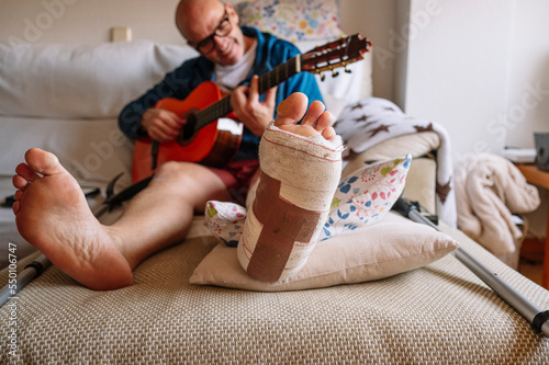 Injured man with bandaged foot and learn to play guitar online.