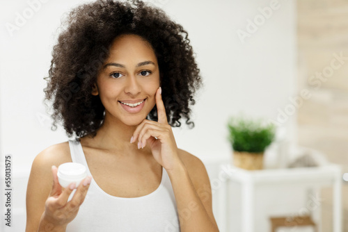 Smiling african american girl applying facial moisturizer while holding jar and looking at camera.
