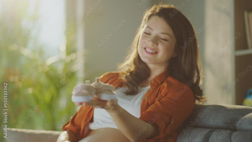 Smiling happy pregnant woman holding baby shoes in her hands while sitting on the sofa at her apartment Smiling happy pregnant woman holding baby shoes in her hands while sitting on the sofa at her apartment