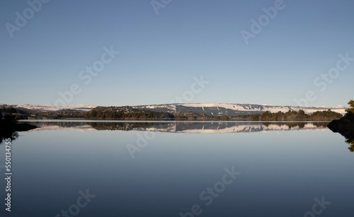 Fototapeta Naklejka Na Ścianę i Meble -  Symmetry in nature. View of the calm lake and shoreline in the horizon. The forest and clear blue sky reflection in the surface of the water.
