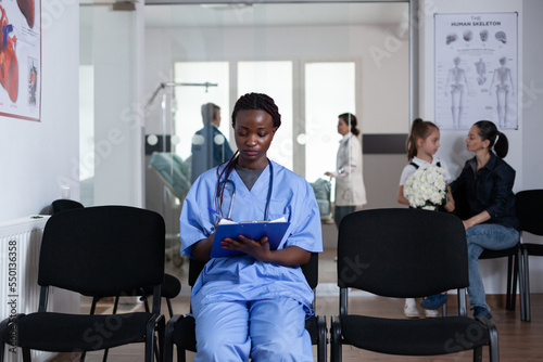 African american nurse filling out patient paperwork sitting at medical tower reception chairs. Young doctor writing prescriptions in hospital waiting room. Medical staff member reviewing patient list