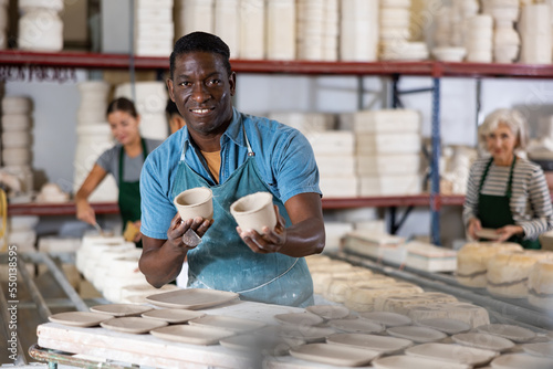Φωτογραφία Positive african-american man potter in apron standing in ceramic workshop, holding crafted cups of bowls, looking at camera and smiling