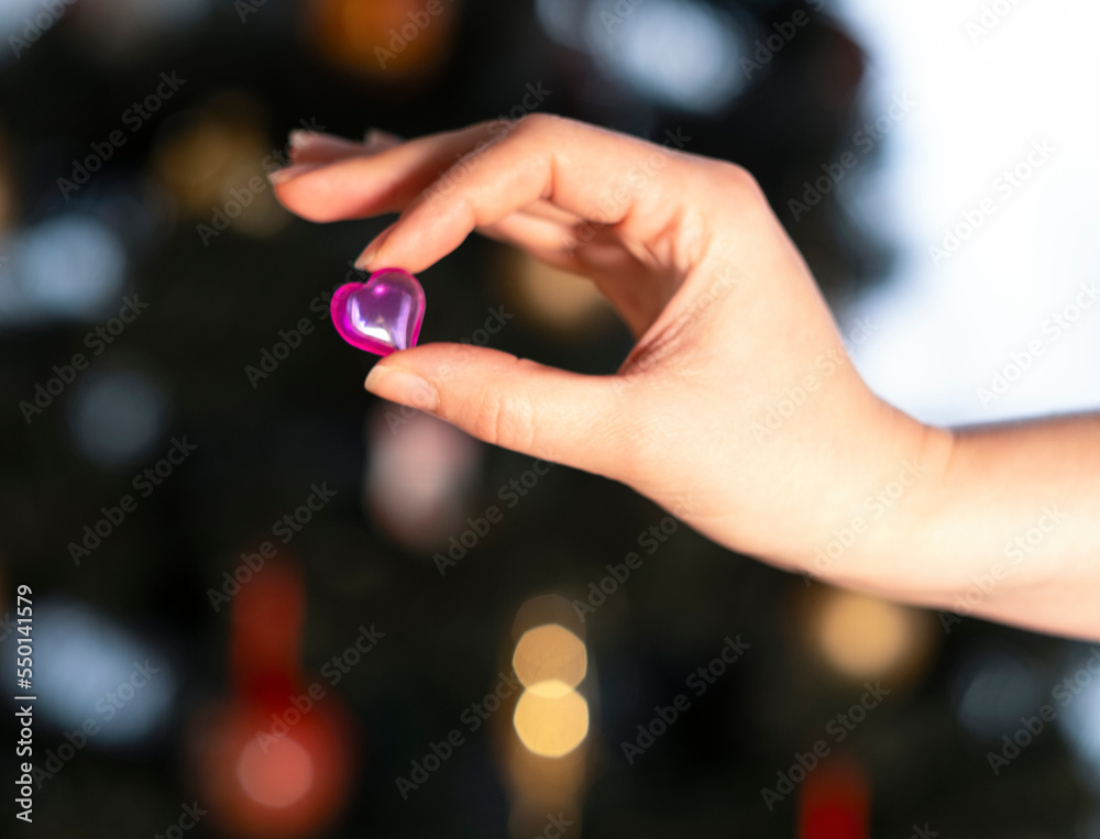 a woman's hand holds a glass pink heart on the background of Christmas lights a woman's hand holds a glass pink heart on the background of Christmas lights
