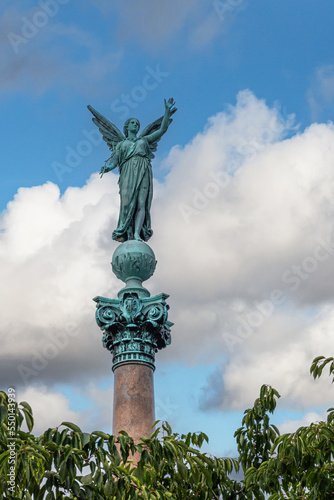 Copenhagen, Denmark - July 24, 2022: Ivar Huitfeldt Column detail. Bronze greenish Victoria statue on top of Admirals brown stone column in Langelinie Park under blue cloudscape photo