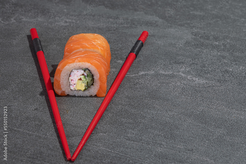 Sushi rolls with salmon, avocado and cucumber with red chopsticks on a black stone (slate) background.
