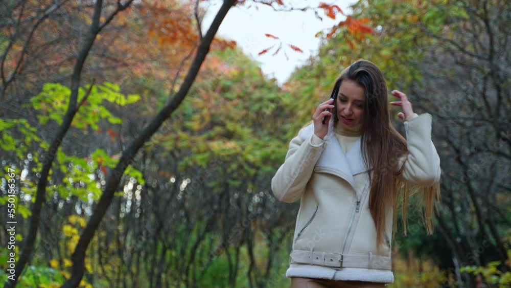 Caucasian lady speaks on the phone walking by the autumn nature. Woman touches her long hair constantly and smiling at conversation. Caucasian lady speaks on the phone walking by the autumn nature. Woman touches her long hair constantly and smiling at conversation.