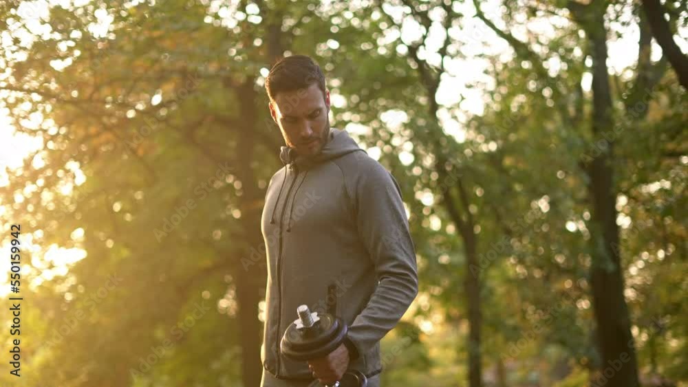 Young handsome athlete doing biceps curls with dumbbells outside in the park.
