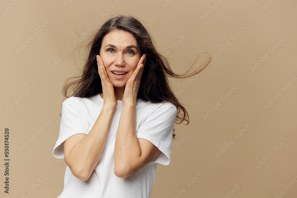 a beautiful, surprised woman with hair flying in the wind stands in a white T-shirt on a beige background, holding her hands near her face a beautiful, surprised woman with hair flying in the wind stands in a white T-shirt on a beige background, holding her hands near her face