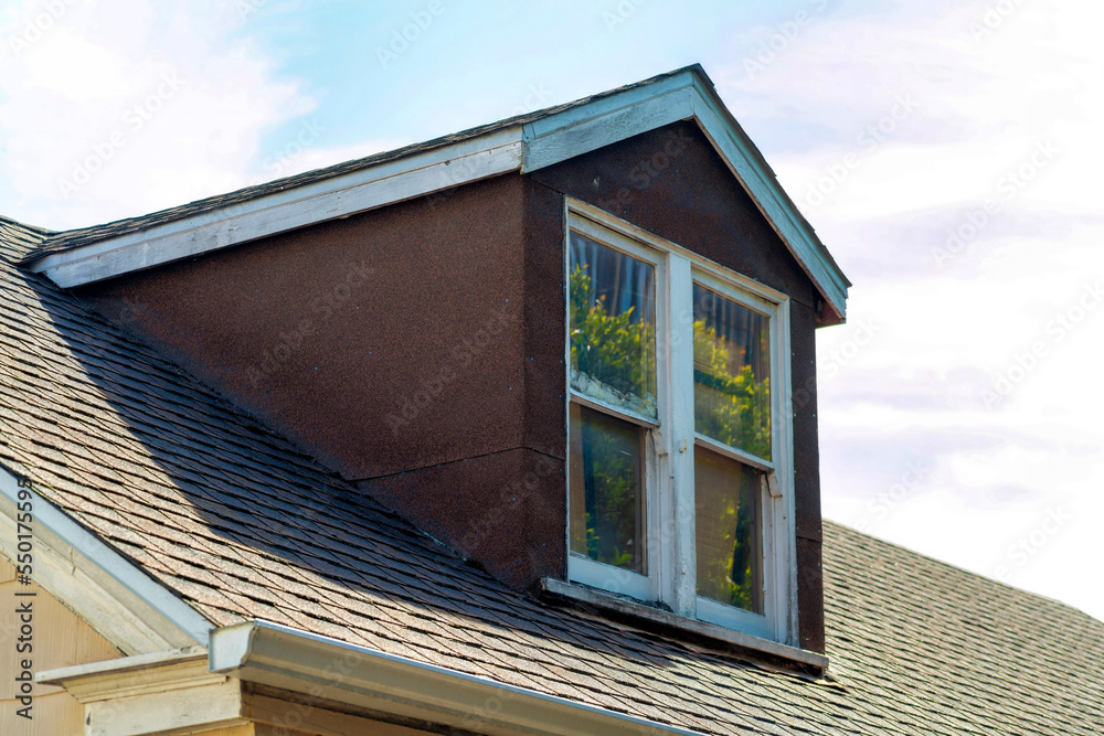 Attic or top floor room window with white trim on edge and on roof in late afternoon shade and sun in the neighborhood Attic or top floor room window with white trim on edge and on roof in late afternoon shade and sun in the neighborhood