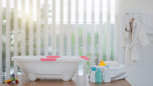 Background of white baby bathtub, towel, shampoo, liquid soap, toys, rubber duck on the table and white gown hanging on the rail. Preparation for taking bath for baby.