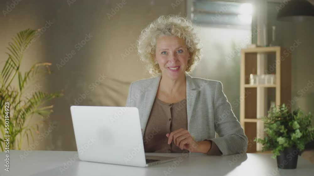 Mature adult curly haired blonde woman sitting at table, typing on her laptop and smiling while looking at camera Mature adult curly haired blonde woman sitting at table, typing on her laptop and smiling while looking at camera