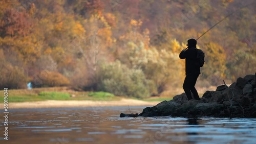 Silhouette of man fishing and throwing a spinning rod into the lake in autumn. View from the lake.
