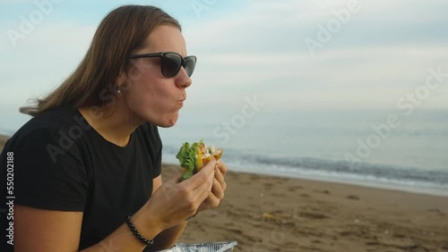 A girl on the seashore, with her mouth wide open, bites off a large piece of burger. Picnic by the sea. photo