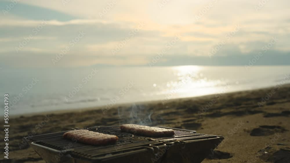 Picnic by the sea at sunset, grilling burger patties. The focus from the sea turns to a close-up of the barbecue. Picnic by the sea at sunset, grilling burger patties. The focus from the sea turns to a close-up of the barbecue.