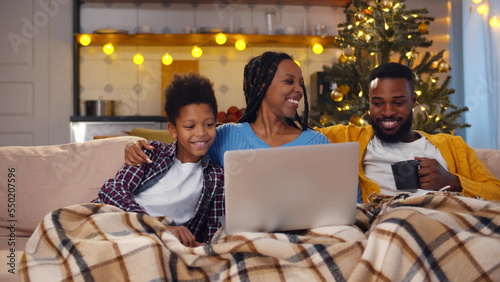 Happy African-American family resting on couch drinking tea and watching movie on laptop on christmas eve