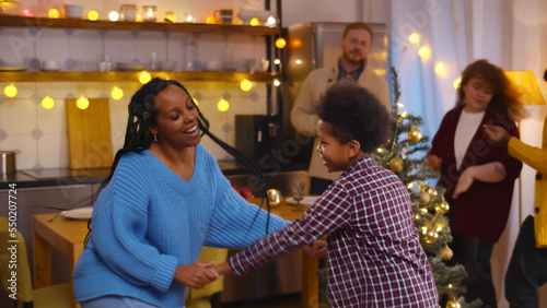 Young African-American woman dancing with son while friends having fun on background celebrating new year