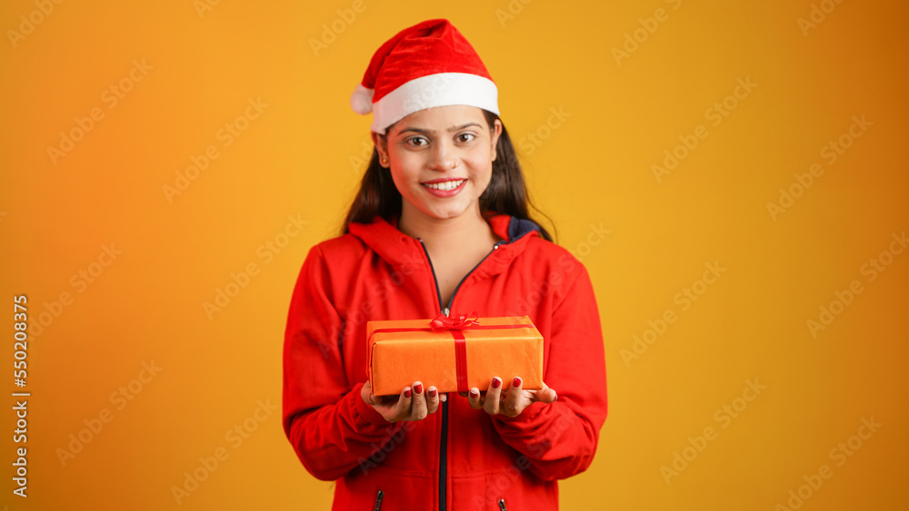 Beautiful young woman with Christmas gift box, Asian Indian girl wearing Santa cap isolated over colour studio background