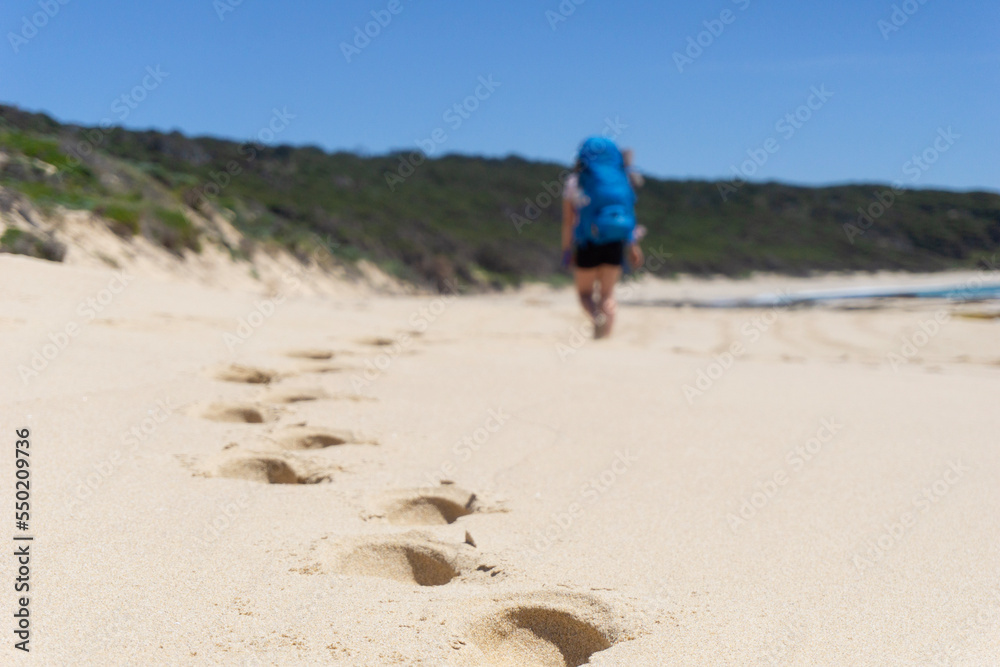 Closeup of footprints on beach while hiker wearing backpack is seen walking along beach with hiking gear in the distance. Sunny day with blue skies along the Cape to Cape track in Western Australia. Closeup of footprints on beach while hiker wearing backpack is seen walking along beach with hiking gear in the distance. Sunny day with blue skies along the Cape to Cape track in Western Australia.