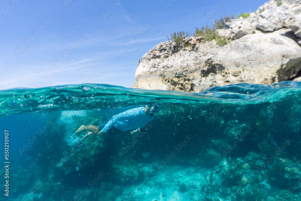 Naklejka premium Underwater split shot of man wearing a rashgaurd swiming in clear aqua blue water alongside rocky cliffside in the Bahamas.