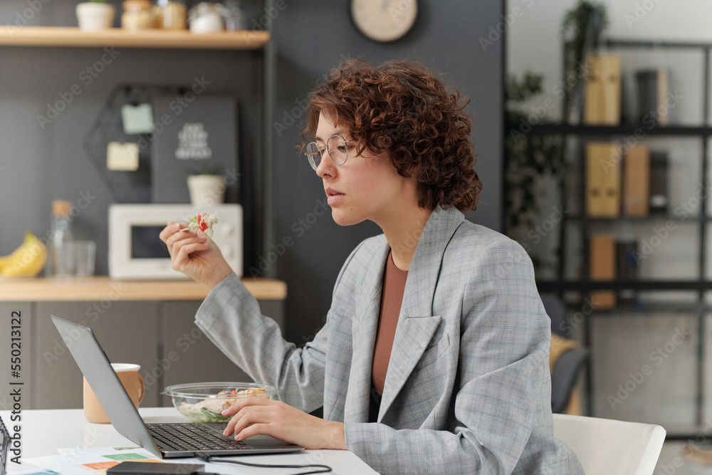 Young businesswoman sitting at table and working on laptop while eating salad during lunch time at office Young businesswoman sitting at table and working on laptop while eating salad during lunch time at office
