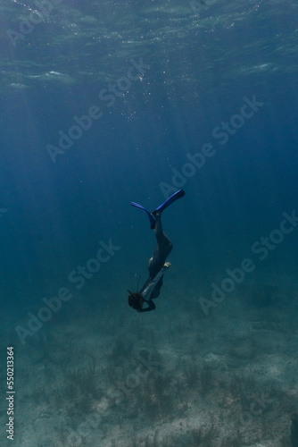 Underwater shot of woman equalizing while freediving down to bottom of shallow ocean in Bahamas with sandy grassy seabed. Sun rays shining through and water is deep blue and moody.